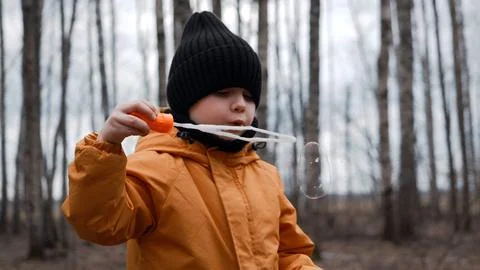 A small child in an orange jacket blows soap bubbles in the forest Stock Photos