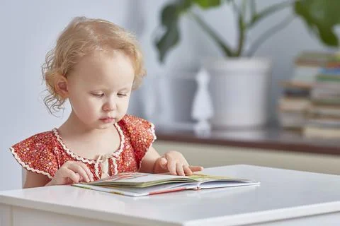 Small child is reading a large book sitting at a table in a bright room. Stock Photos