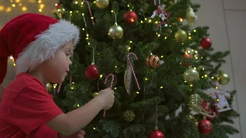 Small child Santa's red hat sits floor and hangs candy cane on Christmas tree Stock Footage 220917408