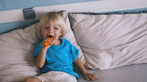 A small child sits on the bed and eats a carrot Stock Footage 220732138