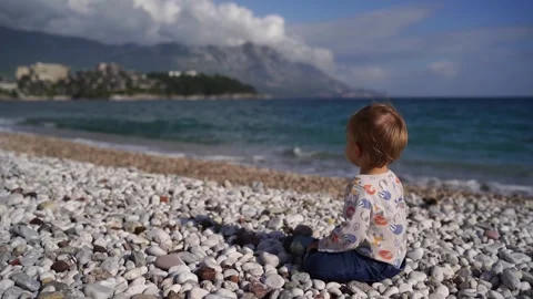 Small child sits bouncing on a pebble beach and points to the sea Stock Footage 256154683