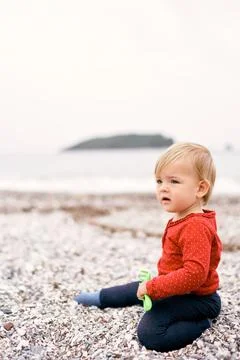 Small child sits on a pebble beach and holds a toy rake in his hand Stock Photos
