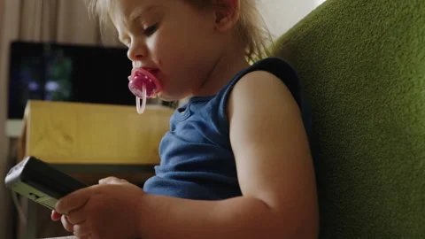 Small child sitting on the green couch sucking a pacifier holding a black game 스톡 동영상 267604505