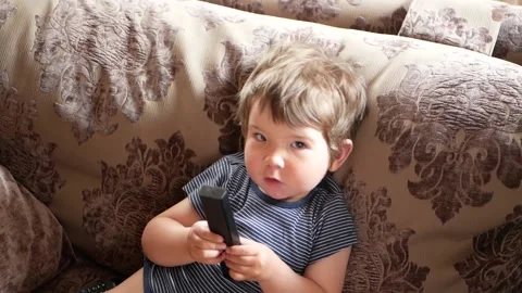 A small child sitting on the sofa, clicks on the TV remote 스톡 동영상 134944262