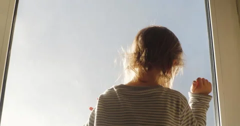 Small child is standing facing the window looking out at the blue sky in rays of Stock Footage 152831715