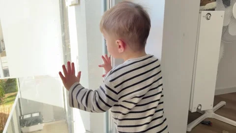 A small child stands by the window in the apartment and looks at the yard Stock Footage 300433679