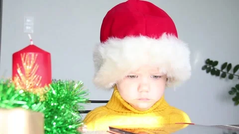 A small child in a yellow sweater and a red Santa Claus hat looks at the tablet Stock Footage 218289959