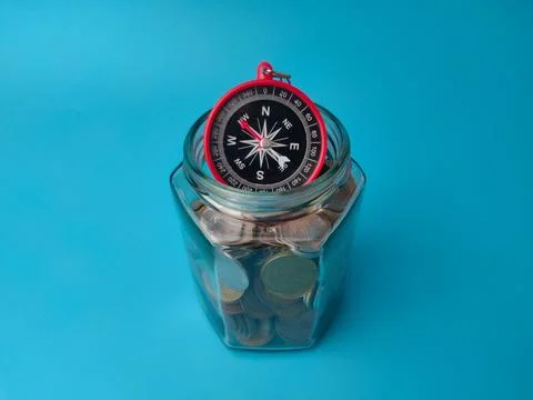 A small compass sits on top of a clear glass jar filled with mixed coins ag.. Stock Photos