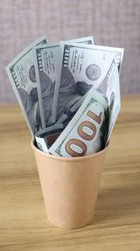 A small cup holds several stacks of hundred dollar bills placed on a wooden Stock Photos