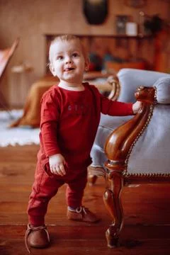 A small cute boy in a red jacket and pants, standing next to achair Stockfoto's