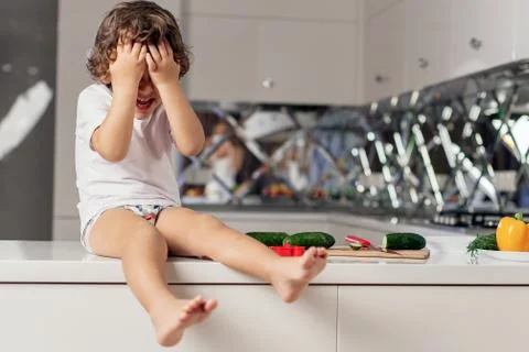 Small cute boy sitting on the kitchen working surface hiding his eyes with his Stock Photos