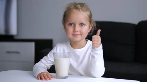 Small cute girl sits at table with glass of milk beside her looking at camera Video stock 277723982