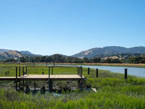 Small dock and deck sitting in a march on a sunny day with river and mountains Stock Photos