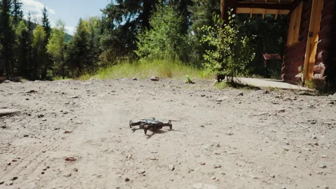 Small drone taking off from dirt ground beside log cabin Video stock 348023907
