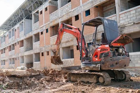A small excavator is parked, digging the soil in front of a building. Stock Photos