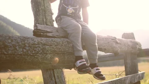 A small fair-haired boy sits on a wooden fence. At sunset in the mountains. Slow Stock Footage 93784715