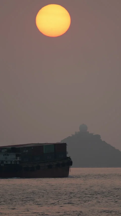 Small feeder container ship sailing at sunset, against island with radar dome Stock Footage 304652872