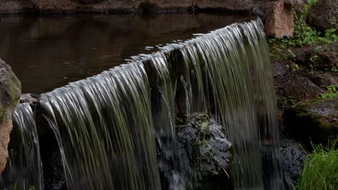 Small garden waterfall flowing over stone ledge into quiet pond with mossy rocks Stock Footage 345453653
