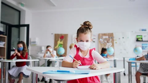 Small girl with face mask sitting on lesson indoors in classroom, looking at Stock Footage