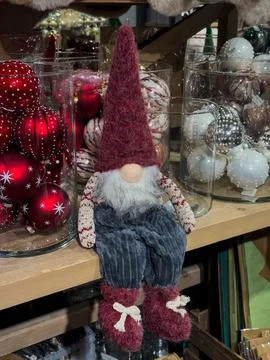A small gnome in a red cap sits on a store window among Christmas balls Stock Photos