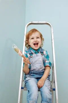 A small happy child sits on a stepladder and holds a brush with paint. Stock Photos