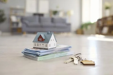 Small House Model on Stack of Money With Keys on Floor of Bright Living Room Stock Photos