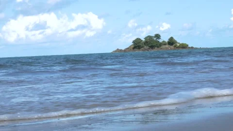 Small island seen from a beach with trees, waves crashing in on sunny day Stock Footage 132172759