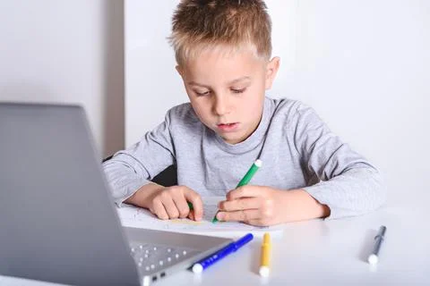 Small kid drawing on a white table with a laptop near. Concept of home school Fotos de archivo