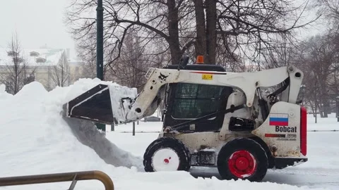 A small loader with a bucket removes snow from a sidewalk in a city park Stock Footage 162315578