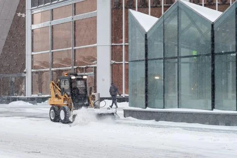 A small loader excavator clears snow from the road along the glass walls Stock Photos