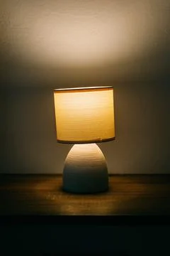 Small modern bedside table lamp glowing with warm ambient light on a wooden.. Stock Photos