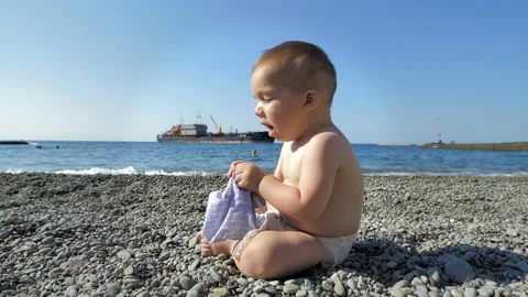 A small one-year-old boy is sitting by the sea on a bright sunny day and playing Stock Footage 159573456
