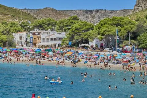 A small pebbly beach in a bay with pine trees on a hot day is full of people Stock Photos