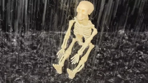 Small plastic skeleton figure sits on a flat surface while water droplets Stock Footage 338172570