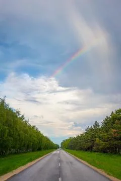 Small rainbow over trees on either side of a straight road Stock Photos