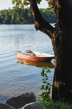 Small red boat floating on a calm lake water, framed by a dark tree trunk o.. Stock Photos