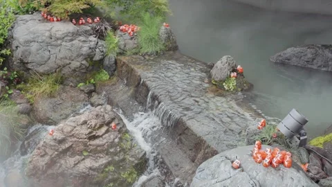 Small red Daruma dolls positioned along mossy rocks beside a gently and foggy Stock Footage 344279202