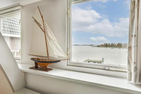 A small sail boat sits on a window sill overlooking Stock Photos