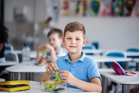 A small school boy sitting at the desk in classroom, eating grapes. Stock Photos