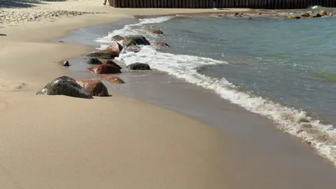 Small sea waves gently break against scattered stones on a sandy beach under a c Видео 310826716