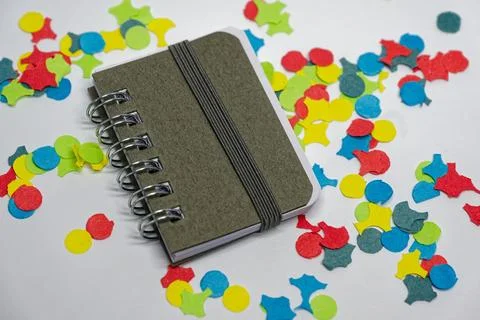 Small spiral notebook on a white surface surrounded by colorful confetti Stock Photos