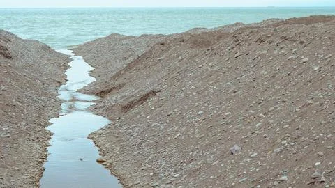 A small stream of water flowing through large piles of pebbles toward the sea Stock Photos