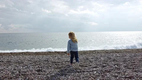 Small three-year-old child run up to cold sea along stone beach in denim jacket Stock Footage 224574130