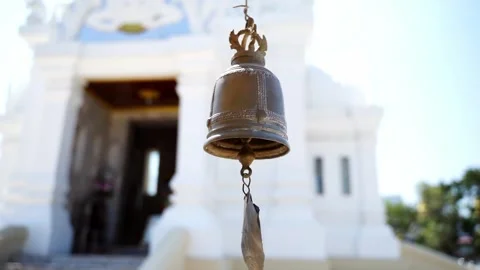 A small traditional bronze temple bell hanging in front of a white Thai Bud.. 動画素材 336982601