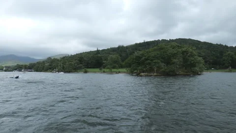 A small, tree-covered island sits on a rippling lake with cloudy hills beyond. Stock Footage 313770780