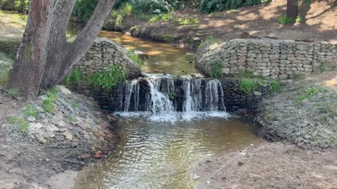 Small waterfall cascading over a stone gabion retaining wall into a calm pond Stock Footage 345550904