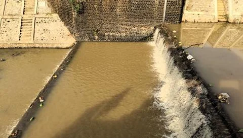 A small waterfall cascading over a stone barrier into muddy water below, wi.. Stock-Fotos