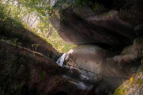 Small waterfall flowing through rocky cave opening in sunlit forest 库存照片