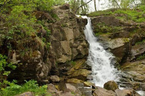 A small waterfall flows in a rapid cascade from a stone wall surrounded by .. Stock Photos
