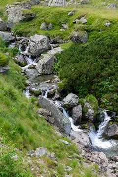 A small waterfall flows through rocks and grass in mountainous terrain Stock Photos